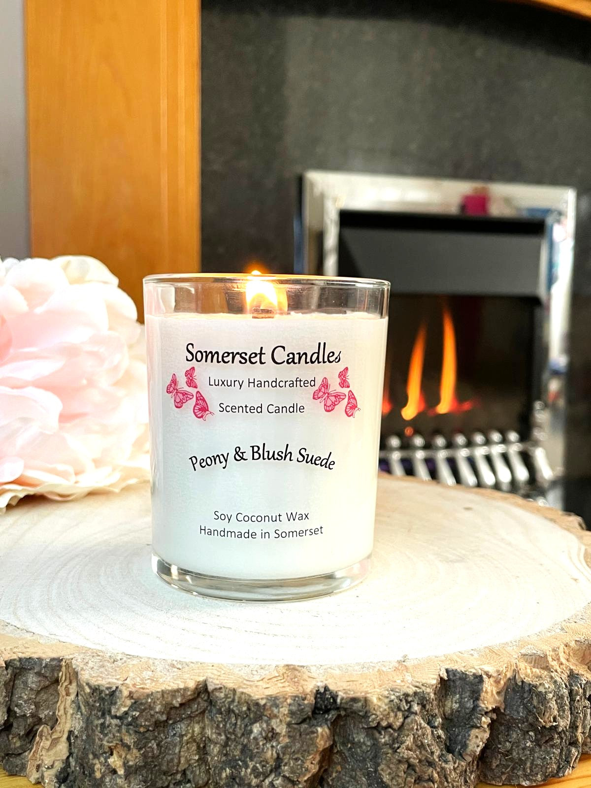 White soy candle in a 7oz clear glass jar with a natural wooden lid. The candle is made from a soy-coconut blend and scented with a soft elegant mix of Peony and Blush Suede. Above the scent name on the label, several pink butterflies are displayed, adding a romantic touch. Clean, minimalist design ideal for gifts, home decor or self-care.