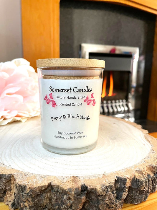 White soy candle in a 7oz clear glass jar with a natural wooden lid. The candle is made from a soy-coconut blend and scented with a soft elegant mix of Peony and Blush Suede. Above the scent name on the label, several pink butterflies are displayed, adding a romantic touch. Clean, minimalist design ideal for gifts, home decor or self-care.