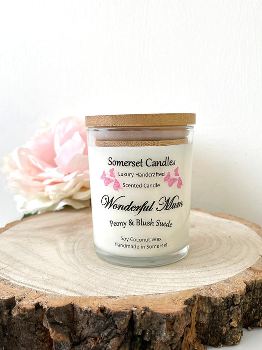White soy candle in a 7oz clear glass jar with a natural wooden lid. The candle is made from a soy-coconut blend and scented with a delicate mix of peony and blush suede. Above the scent name on the label, several pink butterflies are displayed, adding a romantic touch. Clean, minimalist design ideal for gifts, home decor or self-care.