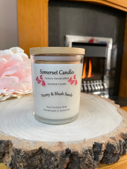 Peony & Blush scented soy candle suede displayed on a wooden board with a beautiful pink peony and a lit fire in the back ground. The candle is white in colour with bright pink butterflies fluttering above the scent name and fitted with a bamboo lid.