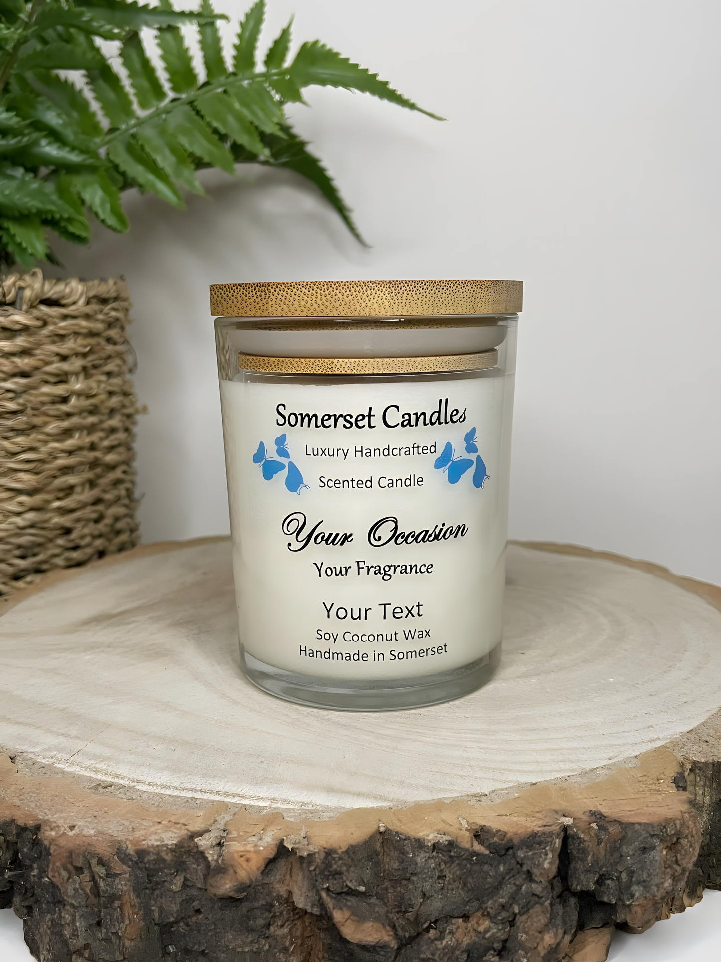 Personalised Scented Soy Candle displayed on a wooden log with a plant behind. The candle is white in colour with blue butterflies above the scent name and a lovely bamboo lid.