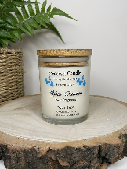 Personalised Scented Soy Candle displayed on a wooden log with a plant behind. The candle is white in colour with blue butterflies above the scent name and a lovely bamboo lid.