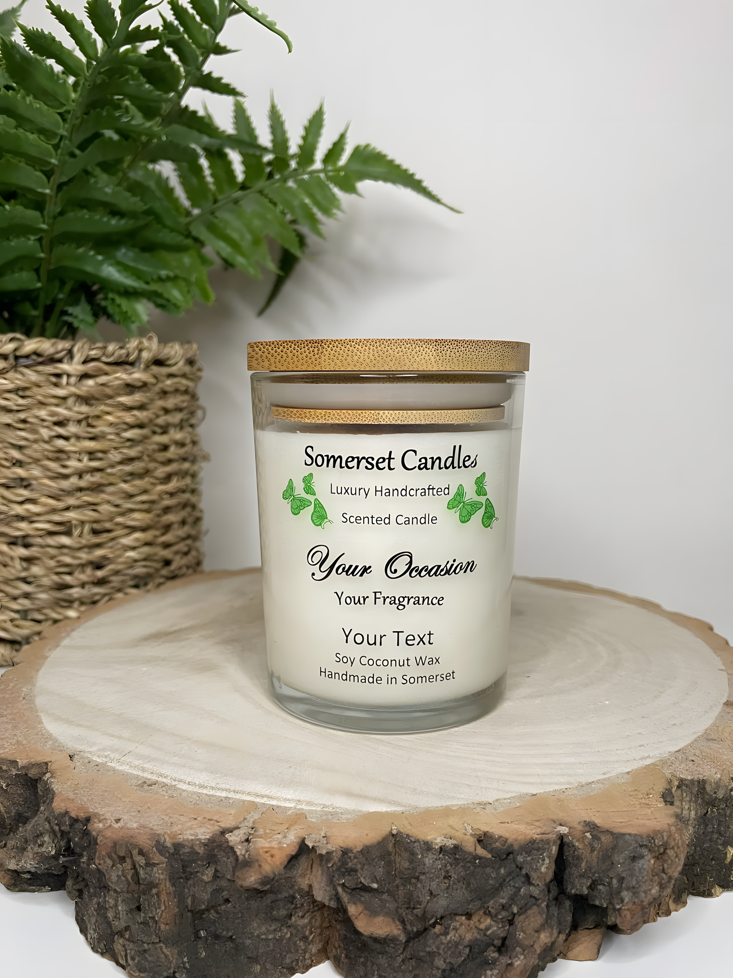 Personalised Scented Soy Candle displayed on a wooden log with a plant behind. The candle is white in colour with green butterflies above the scent name and a lovely bamboo lid.