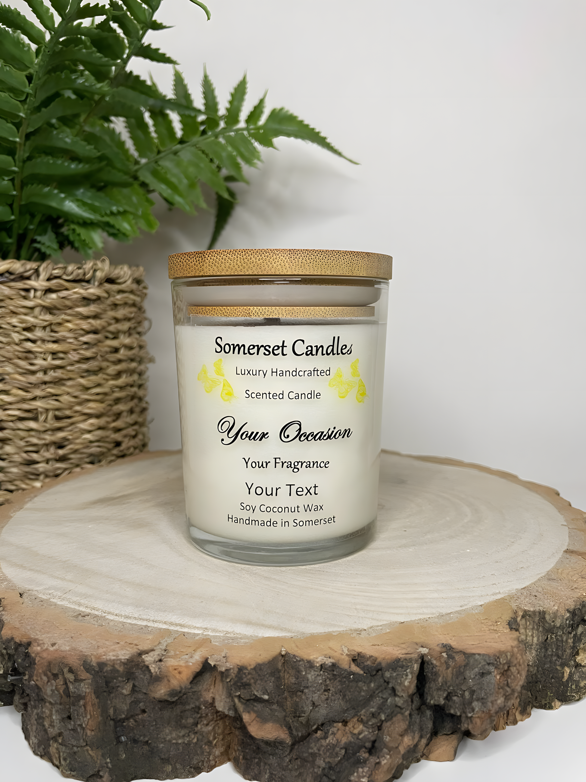 Personalised Scented Soy Candle displayed on a wooden log with a plant behind. The candle is white in colour with yellow butterflies above the scent name and a lovely bamboo lid.