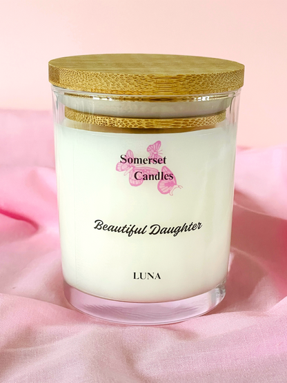 Candle in a glass jar with a wooden lid on a pink fabric background, featuring the brand 'Somerset Candles'.