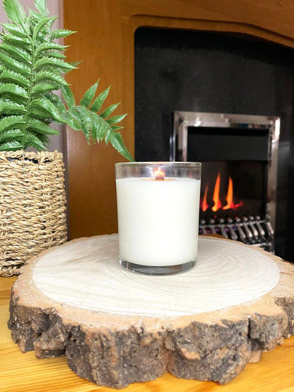 White soy candle in a 7oz clear glass jar with a natural wooden lid. The candle is made from a soy-coconut blend and a variety of fresh fragrances. The candle has no label. The candle is lit in front of a lit fire. Clean, minimalist design ideal for gifts, home decor or self-care.
