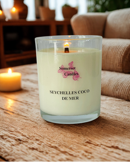A lit Somerset Candles scented candle in a clear glass jar sits on a wooden table in a cozy living room setting, with a blurred sofa and another candle in the background.