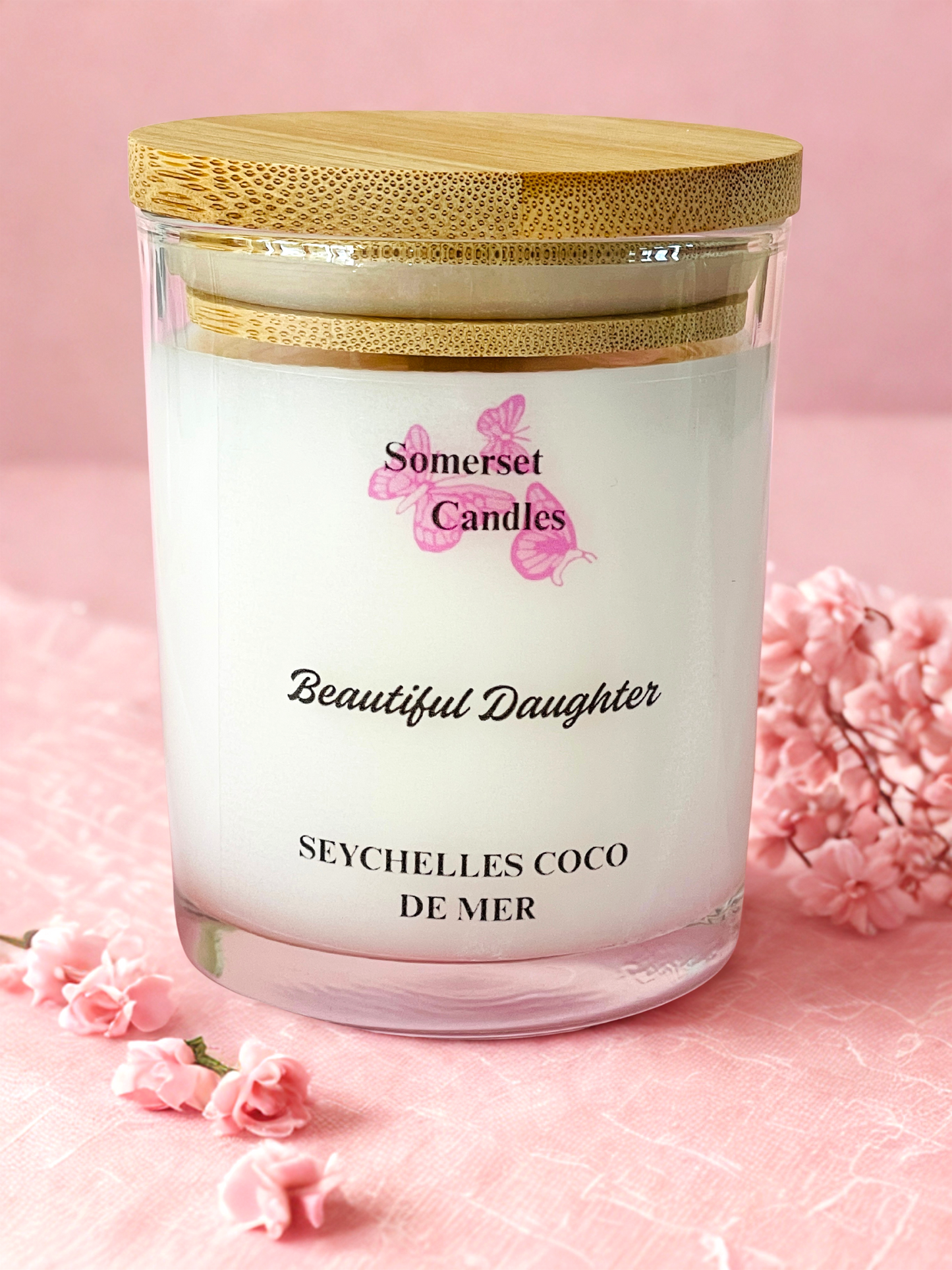 Somerset Candles 'Beautiful Daughter' Seychelles Coco de Mer candle with a wooden lid on a pink background.