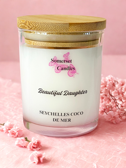 Somerset Candles 'Beautiful Daughter' Seychelles Coco de Mer candle with a wooden lid on a pink background.