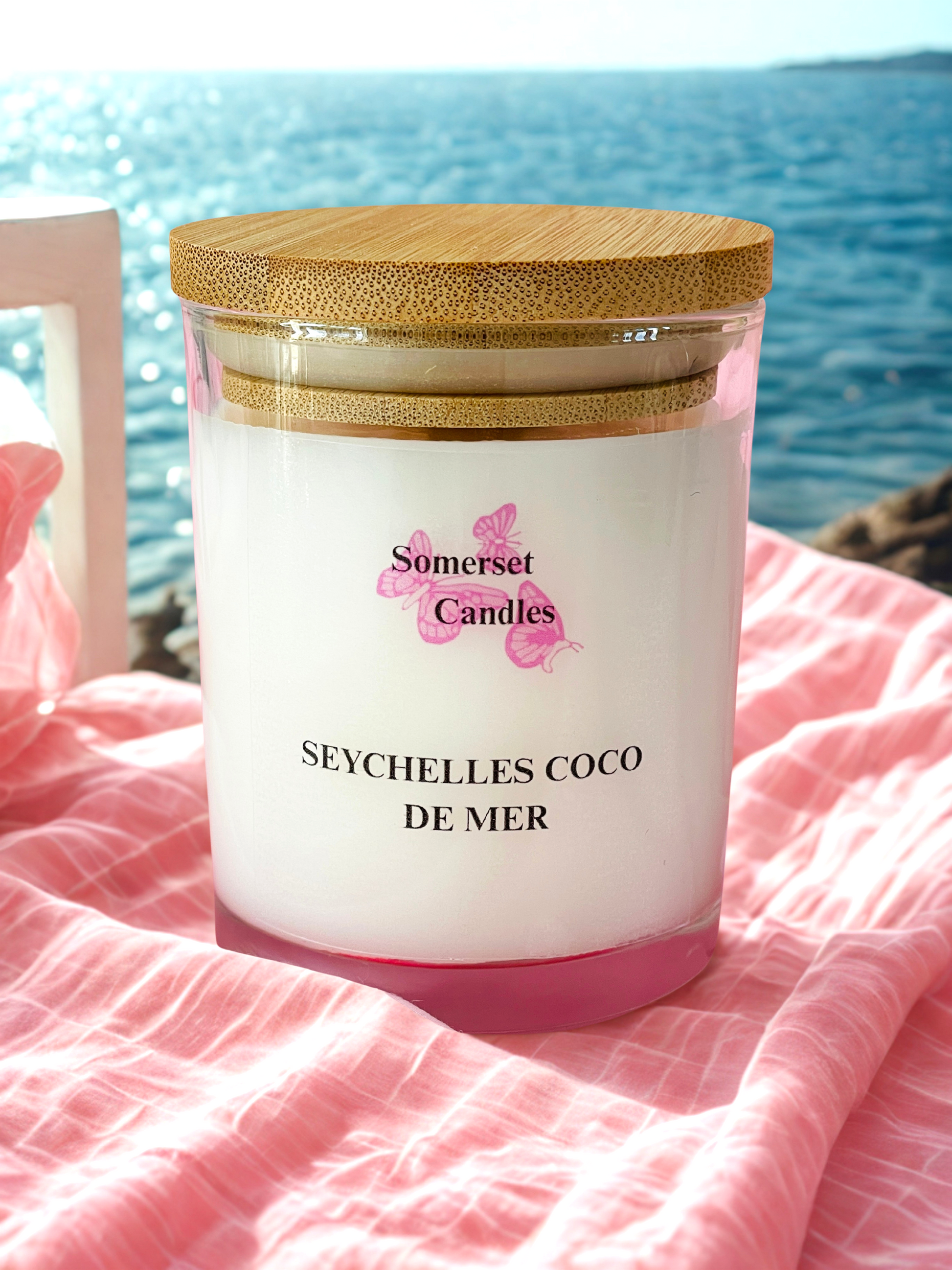 Somerset Candles Seychelles Coco de Mer candle with a wooden lid on a pink blanket by the water.