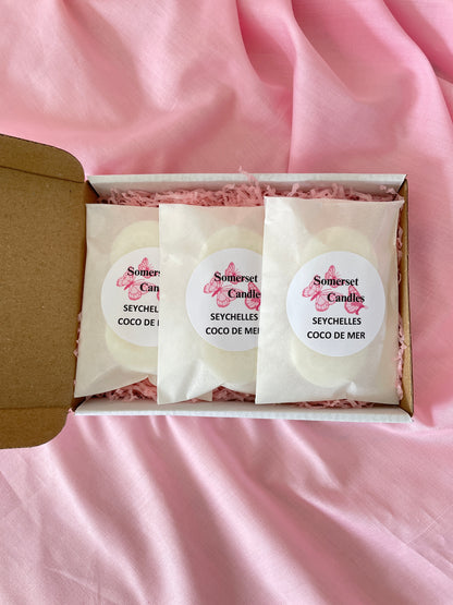 Three individually wrapped Somerset Candles wax melts in a white gift box with pink shredded paper, set against a draped pink fabric background.