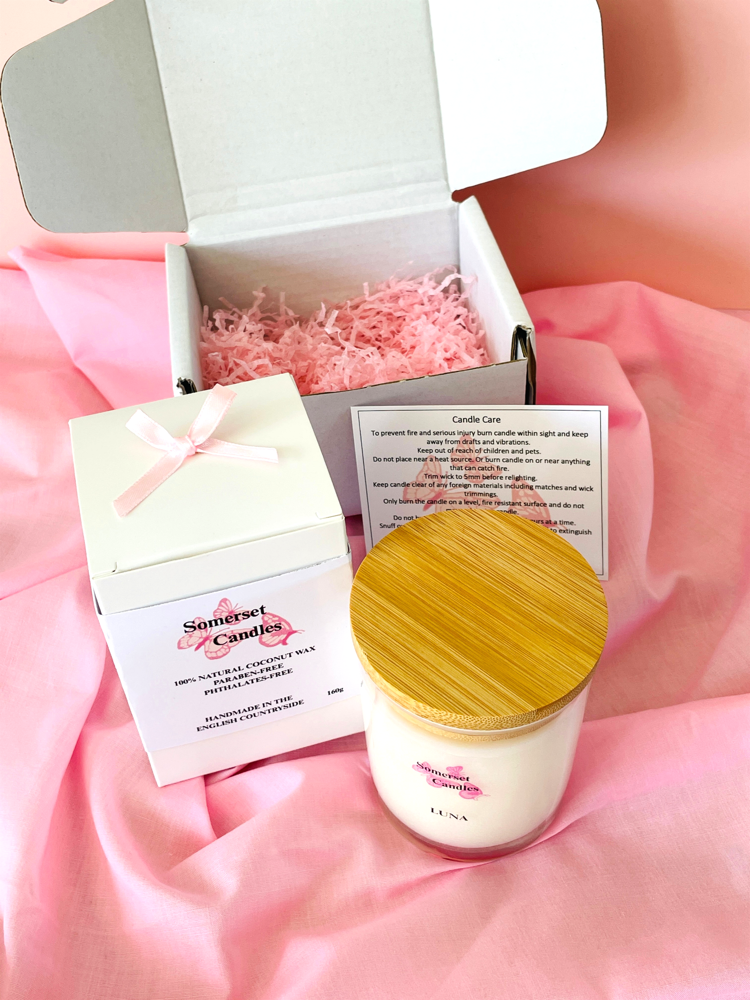 White candle with wooden lid in an open box on a pink fabric background