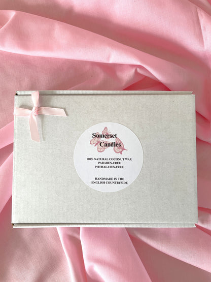 A square light white wax melt box with a pale pink ribbon bow on the left and a round white label reading 'Somerset Candles' in the center, placed on draped pink satin fabric.