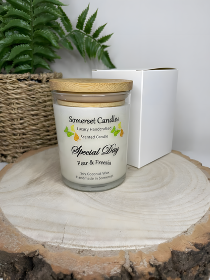 Special Day Pear & Freesia Soy Candle is displayed on a wooden board with a white gift box. The candle is white in colour with bright yellow, green & orange butterflies fluttering above the scent name and fitted with a lovely bamboo lid.