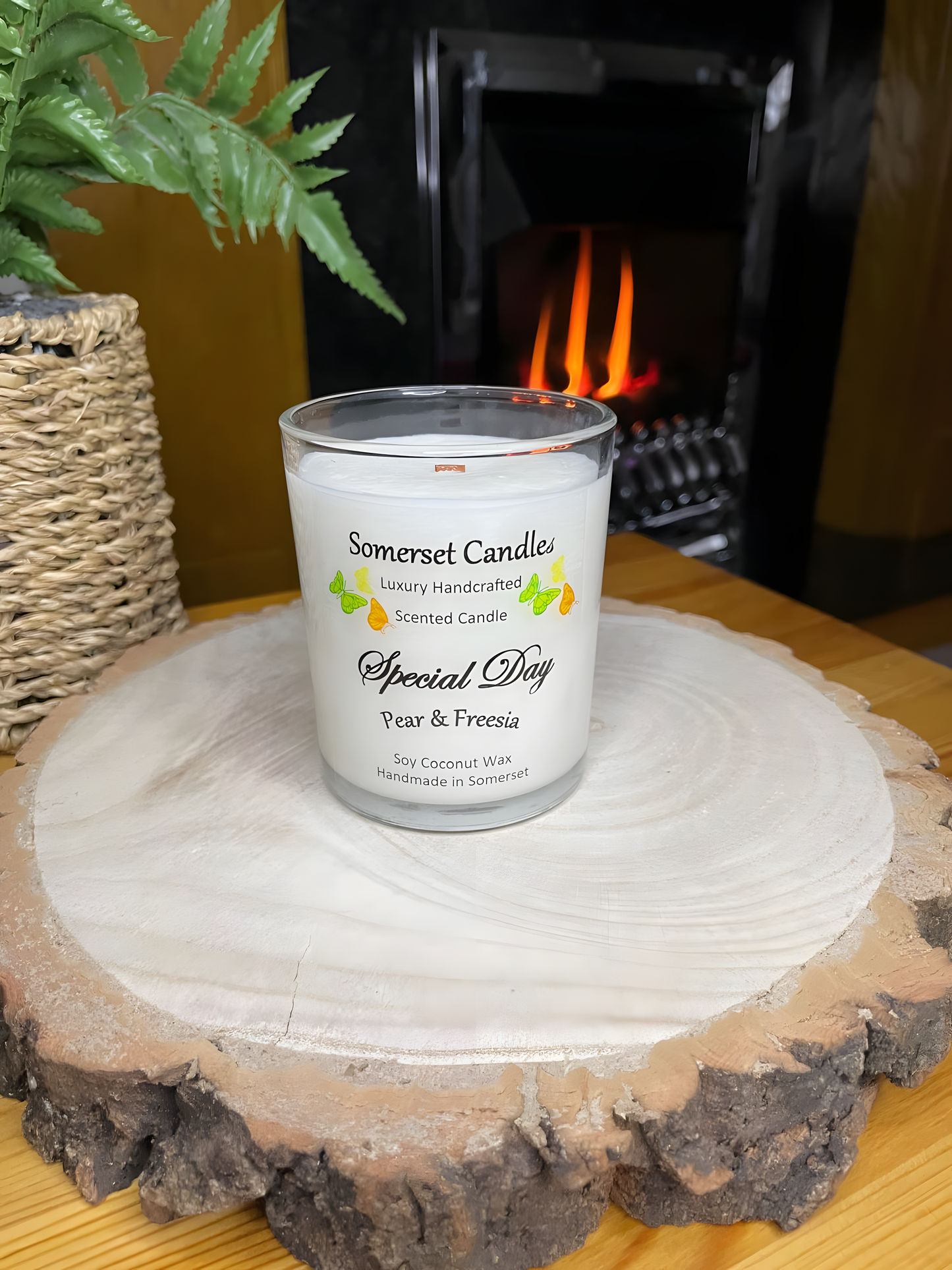 Special Day Pear & Freesia Soy Candle displayed on a wooden board in front of a lit fire. The candle is white in colour with bright yellow, green & orange butterflies fluttering above the scent name and fitted with a lovely bamboo lid.