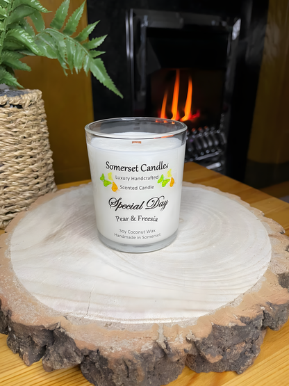 Special Day Pear & Freesia Soy Candle displayed on a wooden board in front of a lit fire. The candle is white in colour with bright yellow, green & orange butterflies fluttering above the scent name and fitted with a lovely bamboo lid.