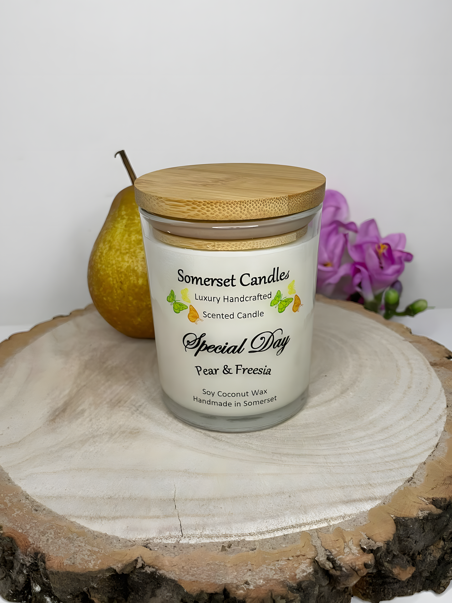 Special Day Pear & Freesia Scented Soy Candle displayed on a wooden board with a fresh pear & freesia. The candle is white in colour with bright yellow, green & orange butterflies fluttering above the scent name and fitted with a lovely bamboo lid.