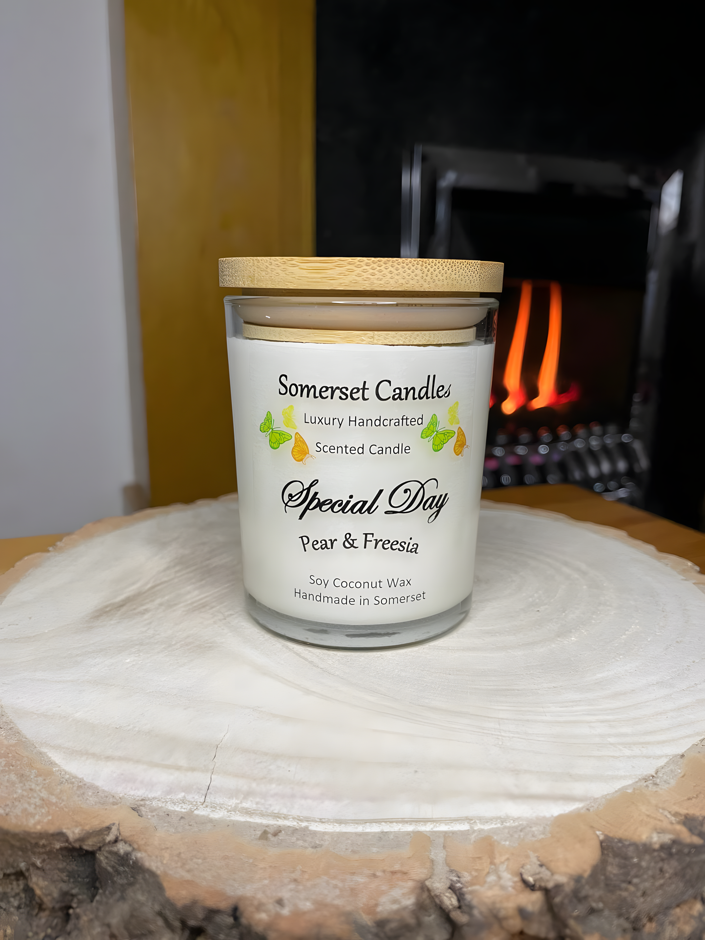 Special Day Pear & Freesia Soy Candle is displayed on a wooden board in front of a lit fire. The candle is white in colour with bright yellow, green & orange butterflies fluttering above the scent name and fitted with a lovely bamboo lid.