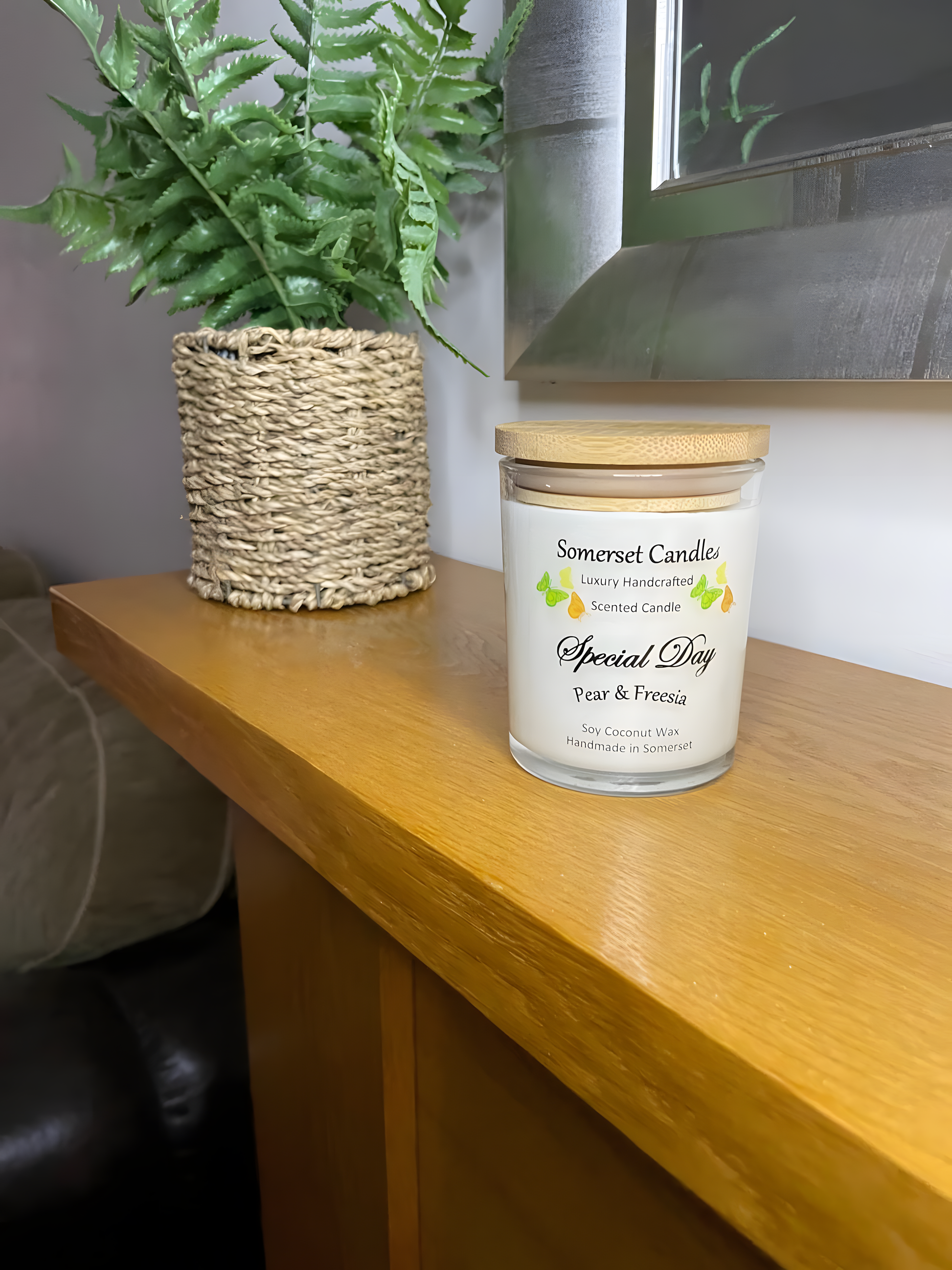 Special Day Pear & Freesia Soy Candle displayed on a the mantelpiece with a plant behind. The candle is white in colour with bright yellow, green & orange butterflies fluttering above the scent name and fitted with a lovely bamboo lid.