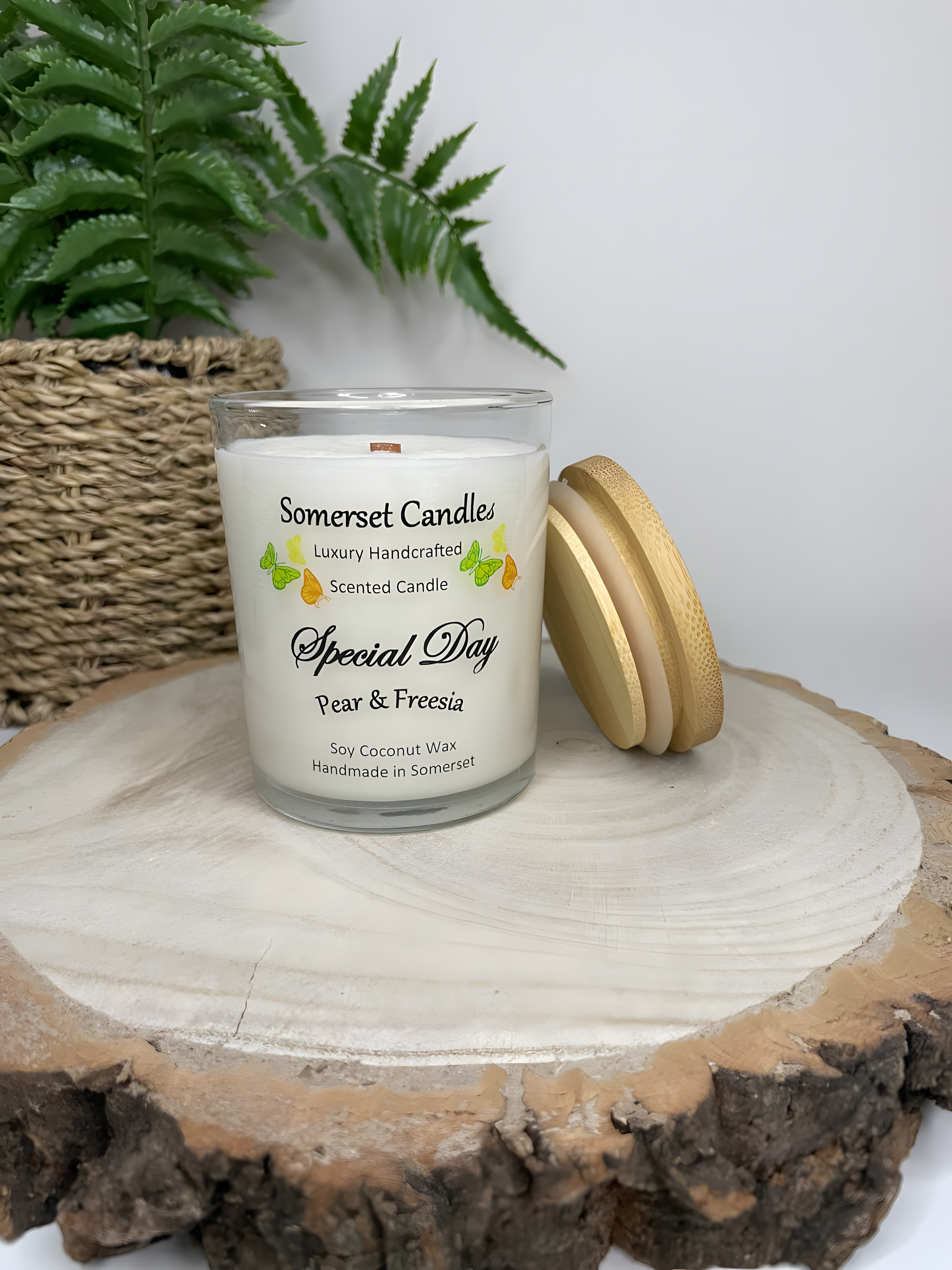 Special Day Pear & Freesia Soy Candle is displayed on a wooden board with a plant behind. The candle is white in colour with bright yellow, green & orange butterflies fluttering above the scent name with a lovely bamboo lid.