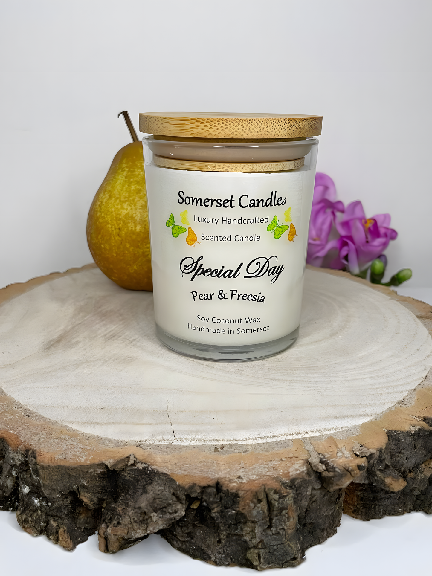 Special Day Pear & Freesia Scented Soy Candle is displayed on a wooden board with a fresh pear & freesia. The candle is white in colour with bright yellow, green & orange butterflies fluttering above the scent name with a lovely bamboo lid.