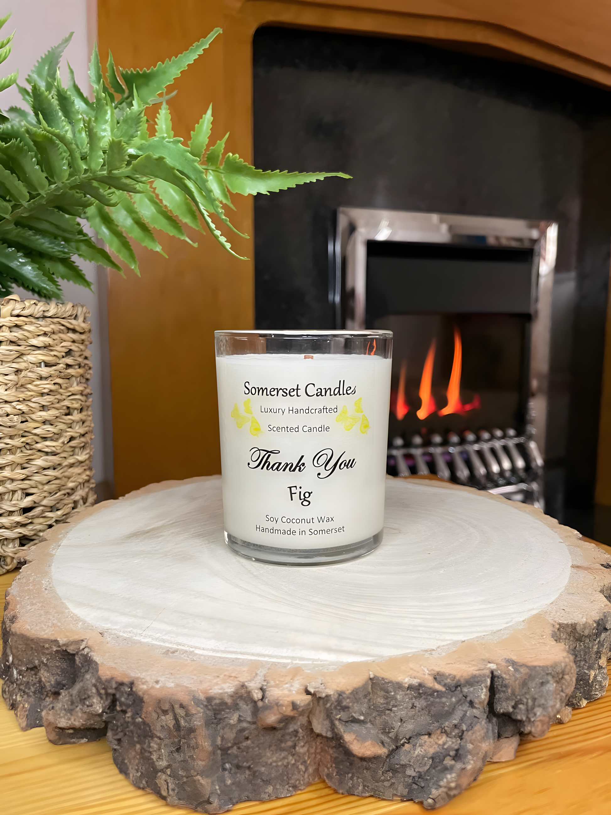 Thank You Fig soy candle is displayed on a wooden board in front of a lit fire. The candle is white in colour with bright yellow butterflies fluttering above the scent name.