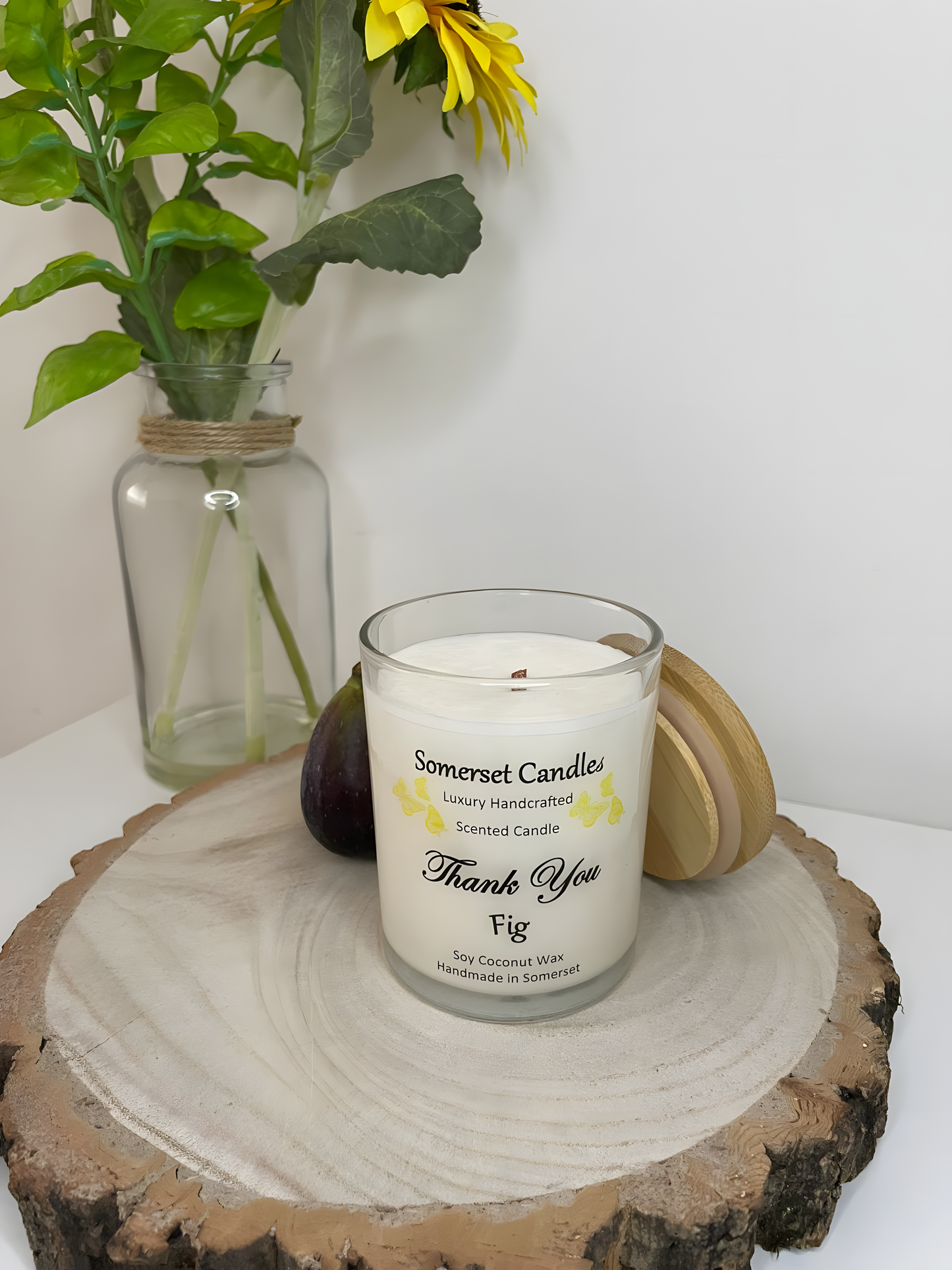 Thank You Fit soy candle is displayed on a wooden board with flowers in the back ground. The candle is white in colour with bright yellow butterflies fluttering above the scent name and fitted with a lovely bamboo lid.