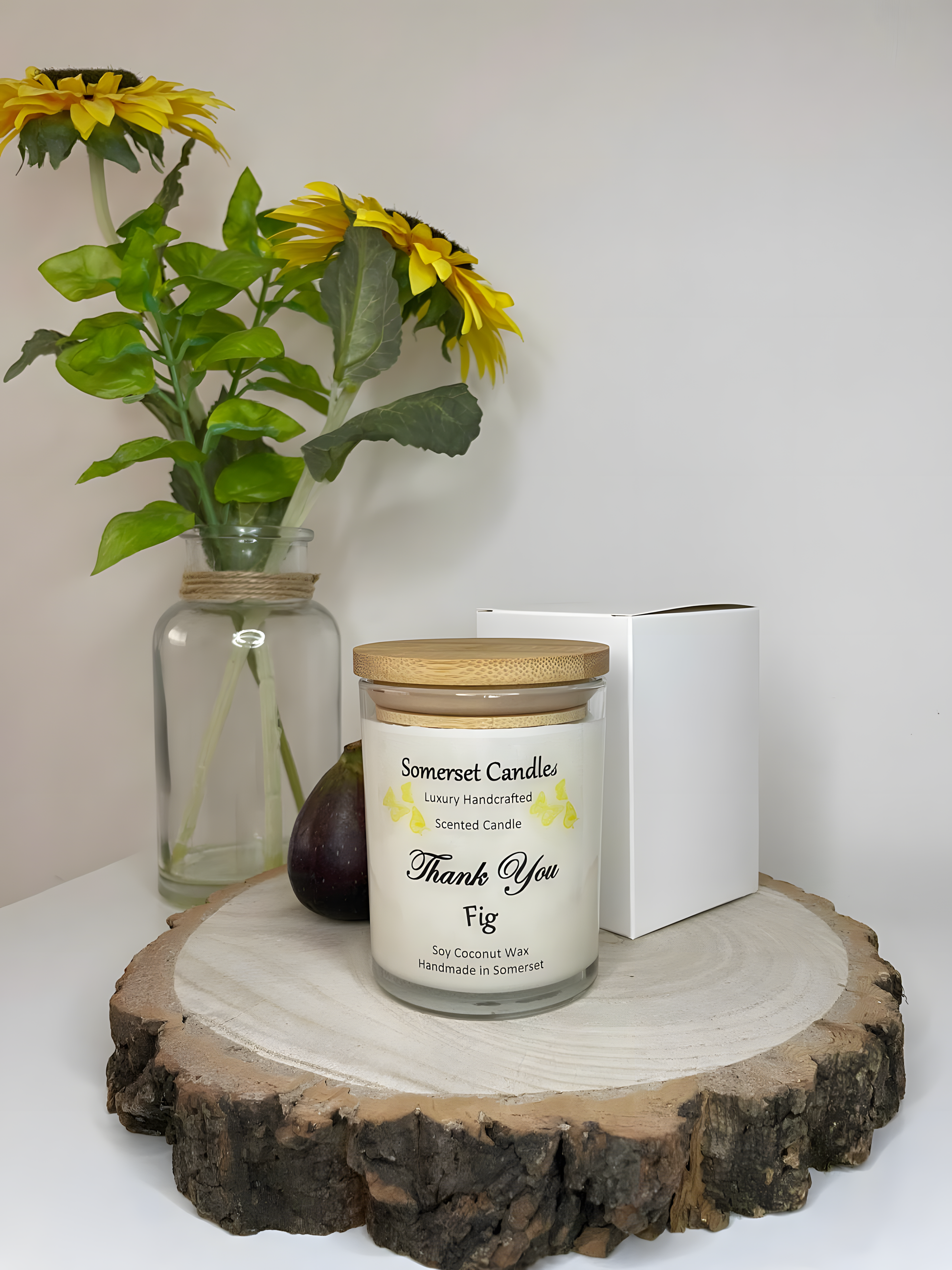 Thank You Fig soy candle is displayed on a wooden board with flowers in the back ground. The candle is white in colour with bright yellow butterflies fluttering above the scent name and fitted with a lovely bamboo lid.