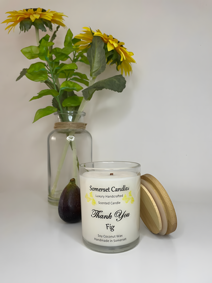 Thank You Fig soy candle is displayed on a white table with flowers in the back ground. The candle is white in colour with bright yellow butterflies fluttering above the scent name and fitted with a lovely bamboo lid.