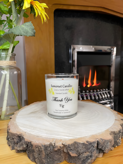 Thank You Fig soy candle is displayed on a wooden board with flowers in front of a lit fire. The candle is white in colour with bright yellow butterflies fluttering above the scent name and fitted with a lovely bamboo lid.