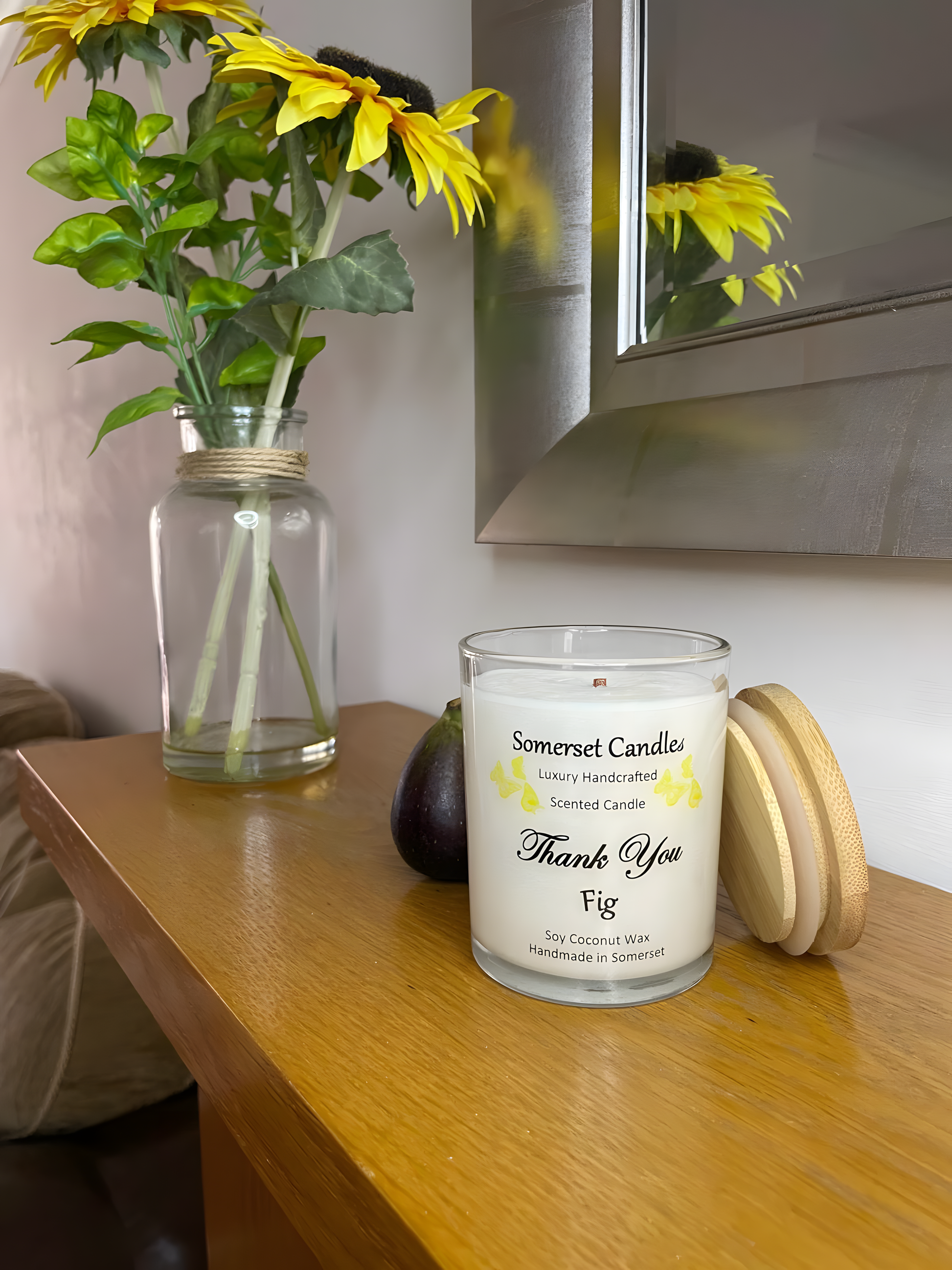 Thank You Fit soy candle is displayed on the mantelpiece with flowers in the back ground. The candle is white in colour with bright yellow butterflies fluttering above the scent name and fitted with a lovely bamboo lid.