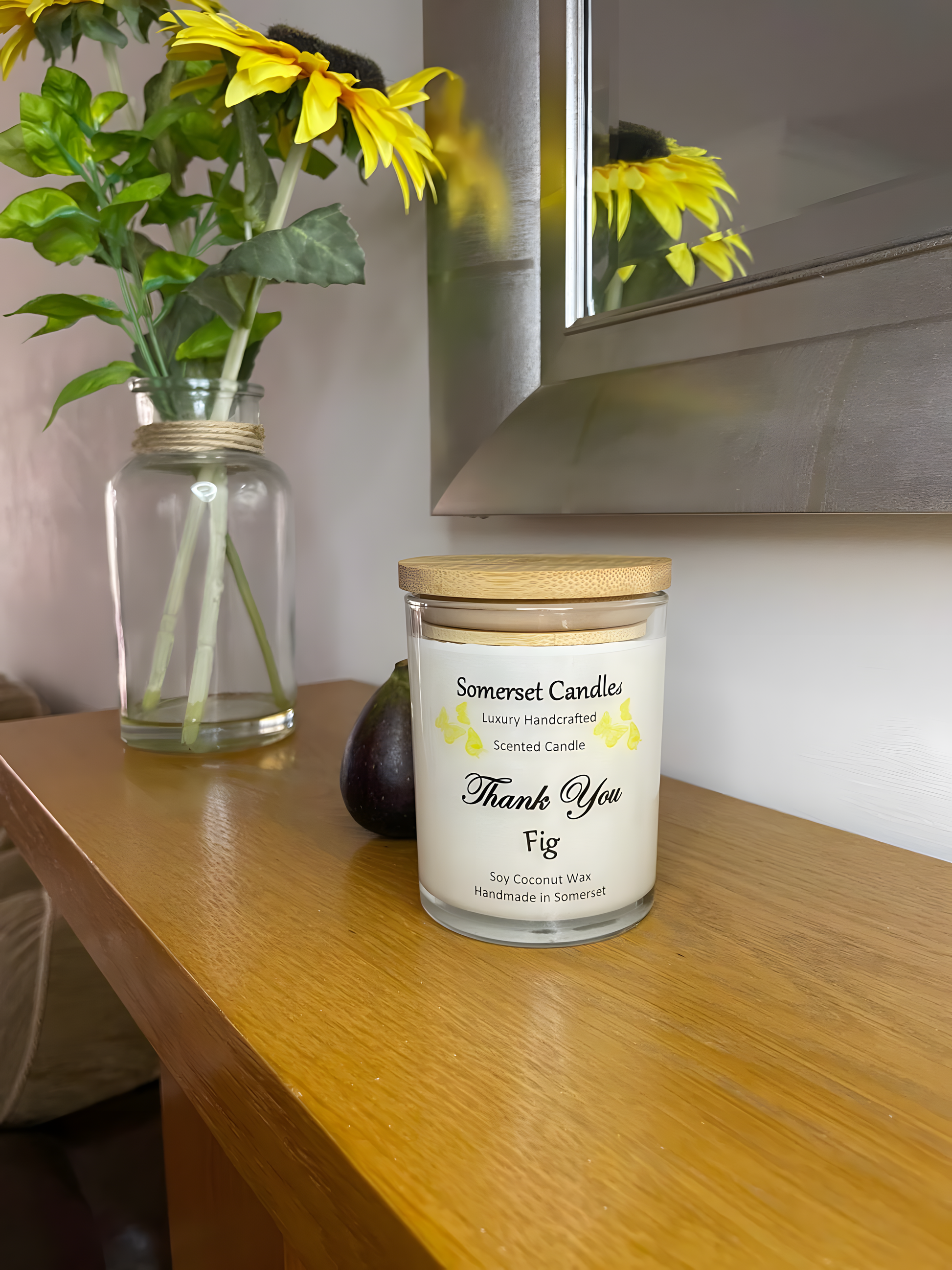 Thank You Fig soy candle is displayed on the mantelpiece with flowers in the back ground. The candle is white in colour with bright yellow butterflies fluttering above the scent name and fitted with a lovely bamboo lid.