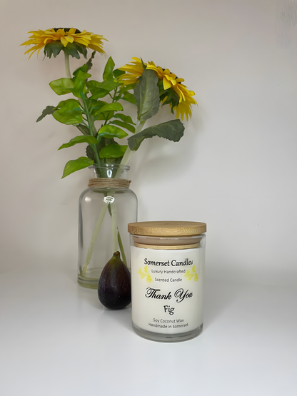 Thank You Fig soy candle is displayed on a white table with flowers in the back ground. The candle is white in colour with bright yellow butterflies fluttering above the scent name and fitted with a lovely bamboo lid.