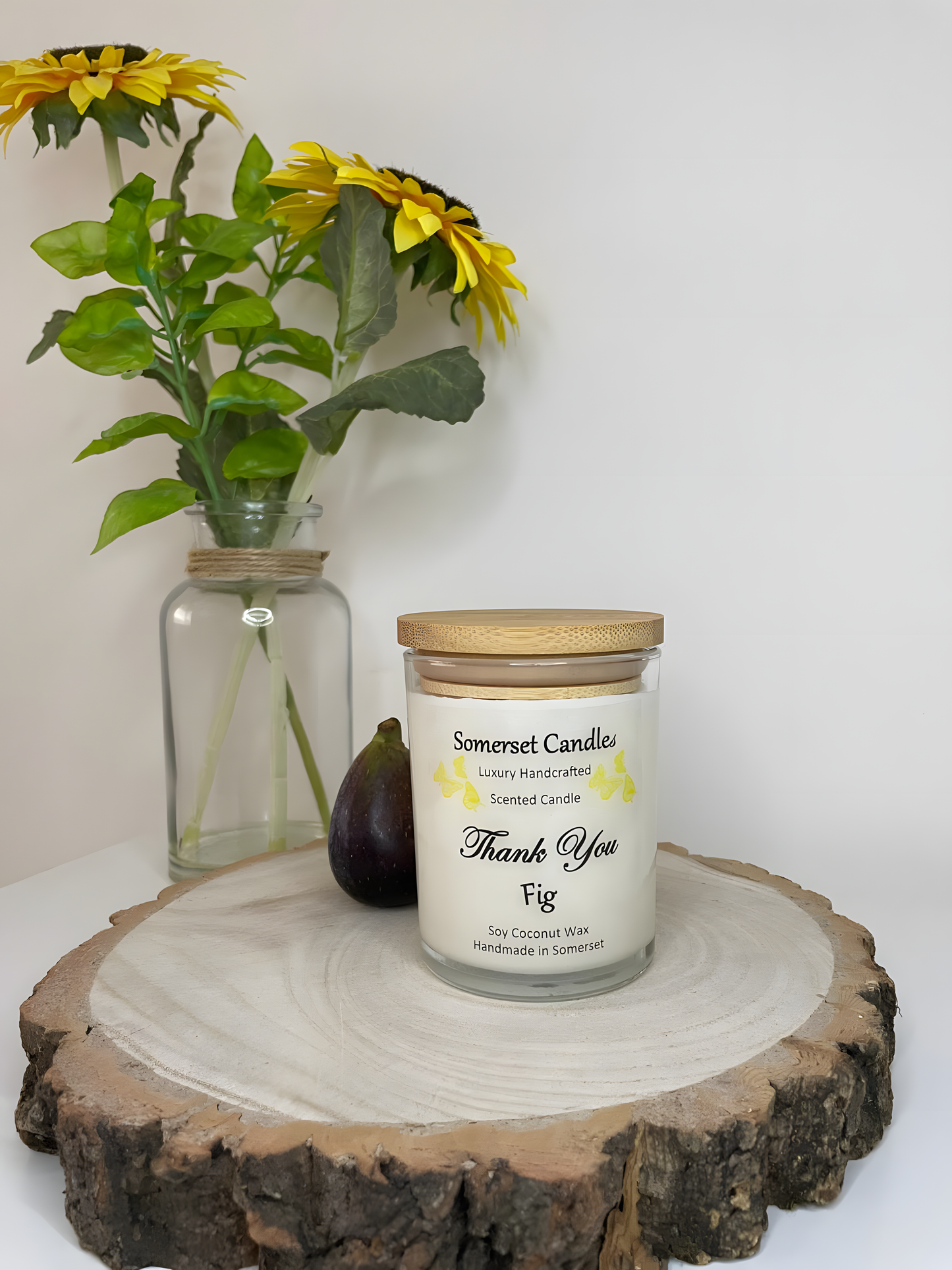 Thank You Fig soy candle is displayed on a wooden board with flowers in the back ground. The candle is white in colour with bright yellow butterflies fluttering above the scent name and fitted with a lovely bamboo lid.