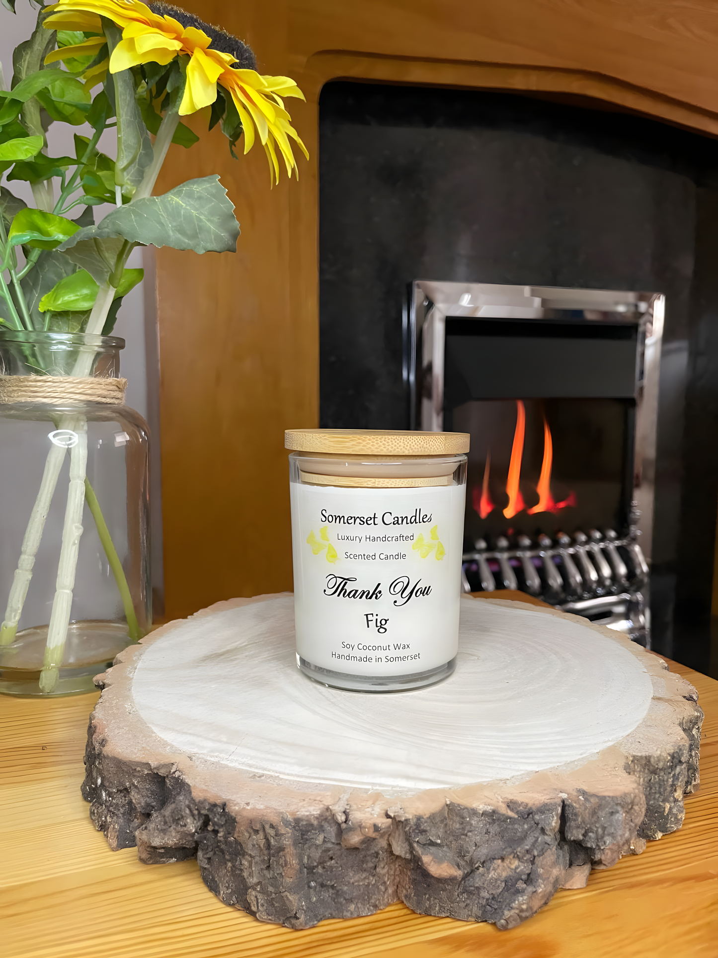 Thank You Fit soy candle is displayed on a wooden board with flowers in front of a lit fire. The candle is white in colour with bright yellow butterflies fluttering above the scent name and fitted with a lovely bamboo lid.