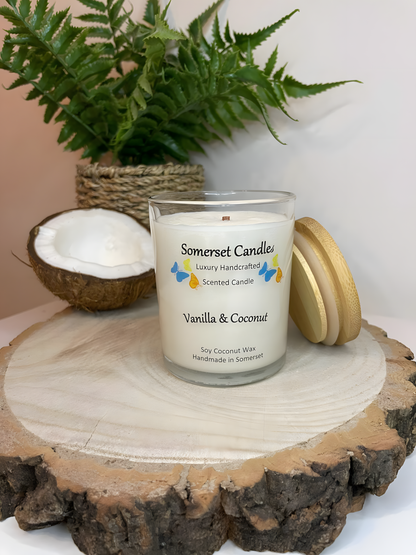 Vanilla & Coconut Scented Soy Candle displayed on a wooden board with half a coconut. The candle is white in colour with bright blue, yellow & orange butterflies fluttering above the scent name and with a lovely bamboo lid.