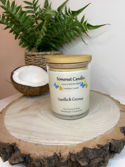 Vanilla & Coconut Scented soy candle displayed on a wooden board with half a coconut. The candle is white in colour with bright blue, yellow & orange butterflies fluttering above the scent name and fitted with a lovely bamboo lid.