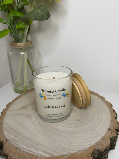 Vanilla & Coconut Scented Soy Candle displayed on a wooden board. The candle is white in colour with bright blue, yellow & orange butterflies fluttering above the scent name. There is lid lent against the side of the candle and the wick can be seen.