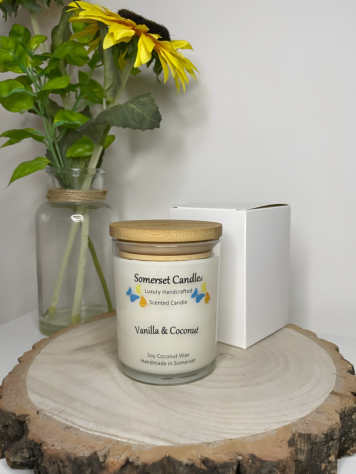 Vanilla & Coconut Scented Soy Candle displayed on a wooden board with half a coconut and gift box. The candle is white in colour with bright blue, yellow & orange butterflies fluttering above the scent name and fitted with a lovely bamboo lid.