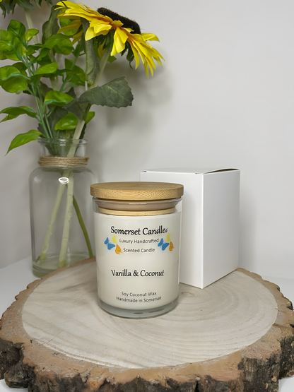 Vanilla & Coconut Scented Soy Candle displayed on a wooden board with half a coconut and gift box. The candle is white in colour with bright blue, yellow & orange butterflies fluttering above the scent name and fitted with a lovely bamboo lid.