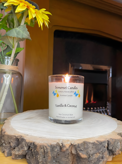 Vanilla & Coconut Scented Soy Candle displayed on a wooden board in front of a fire. The candle is white in colour with bright blue, yellow & orange butterflies fluttering above the scent name. The candle has a clear glass container and is lit.