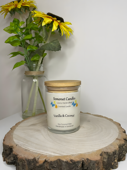 Vanilla & Coconut Scented Soy Candle displayed on a wooden board. The candle is white in colour with bright blue, yellow & orange butterflies fluttering above the scent name and fitted with a lovely bamboo lid. The candle has a clear glass container.