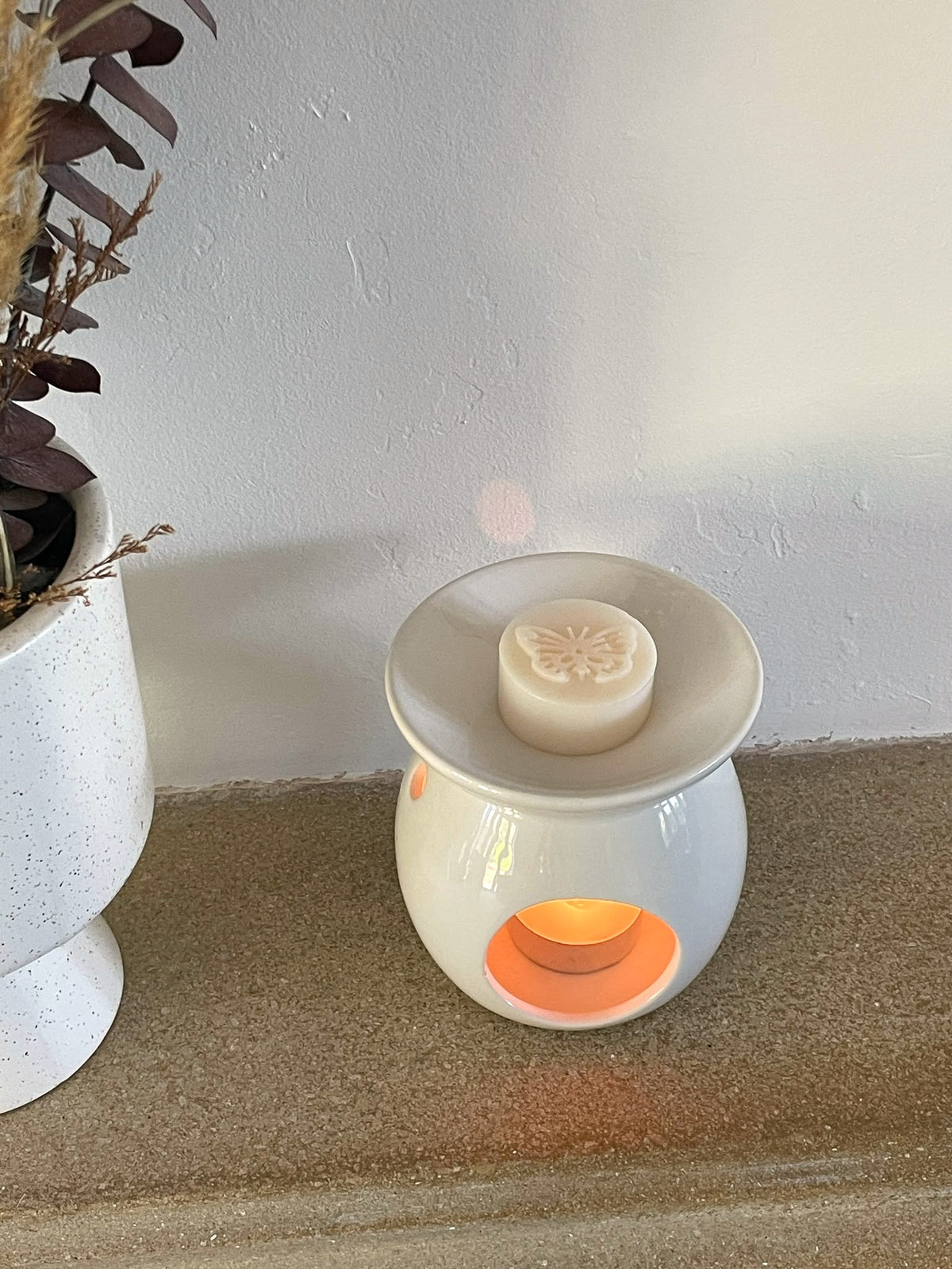 A white ceramic wax warmer with a lit tea light candle inside, shown on a countertop next to a white speckled planter against a light wall.