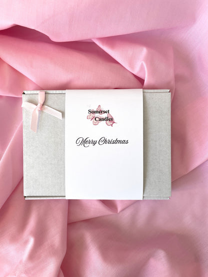 A square gift box wrapped with a white paper band reading 'Somerset Candles' and 'Happy Birthday,' tied with a pale pink ribbon, placed on draped pink fabric.
