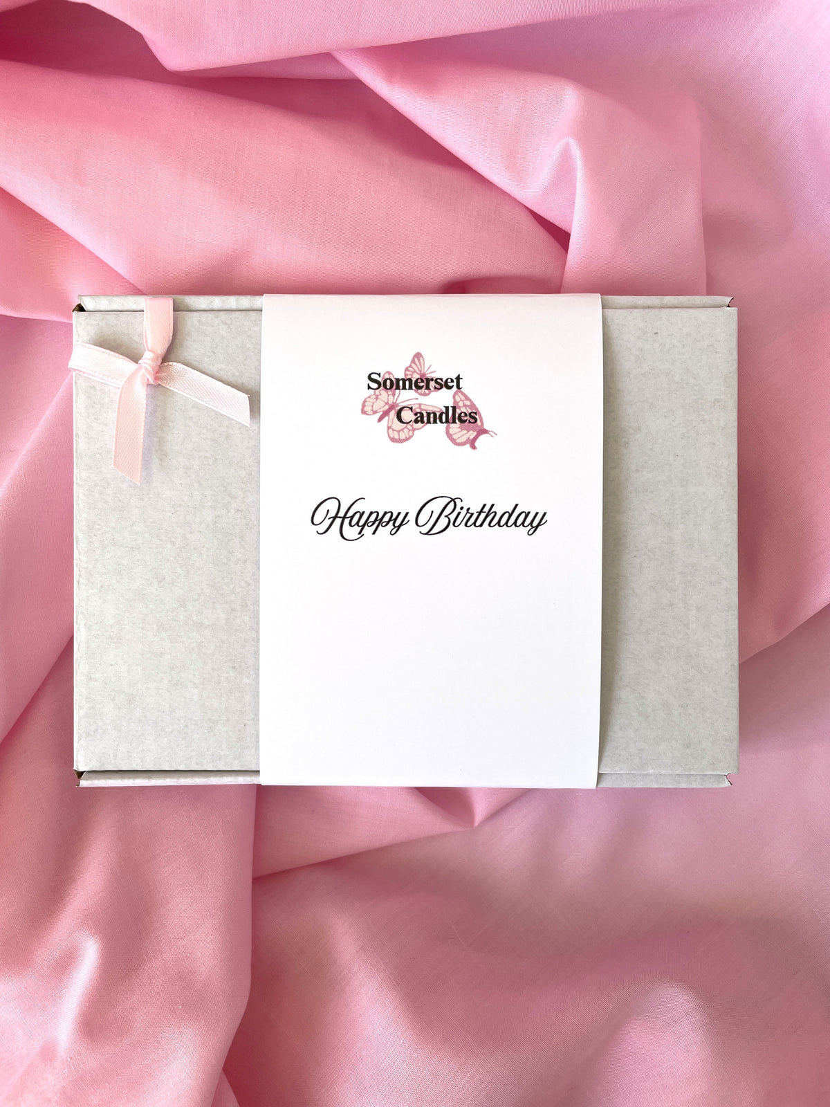 A square gift box wrapped in a white card band reading 'Somerset Candles Happy Birthday' with a small pink butterfly graphic, placed on draped pink fabric.
