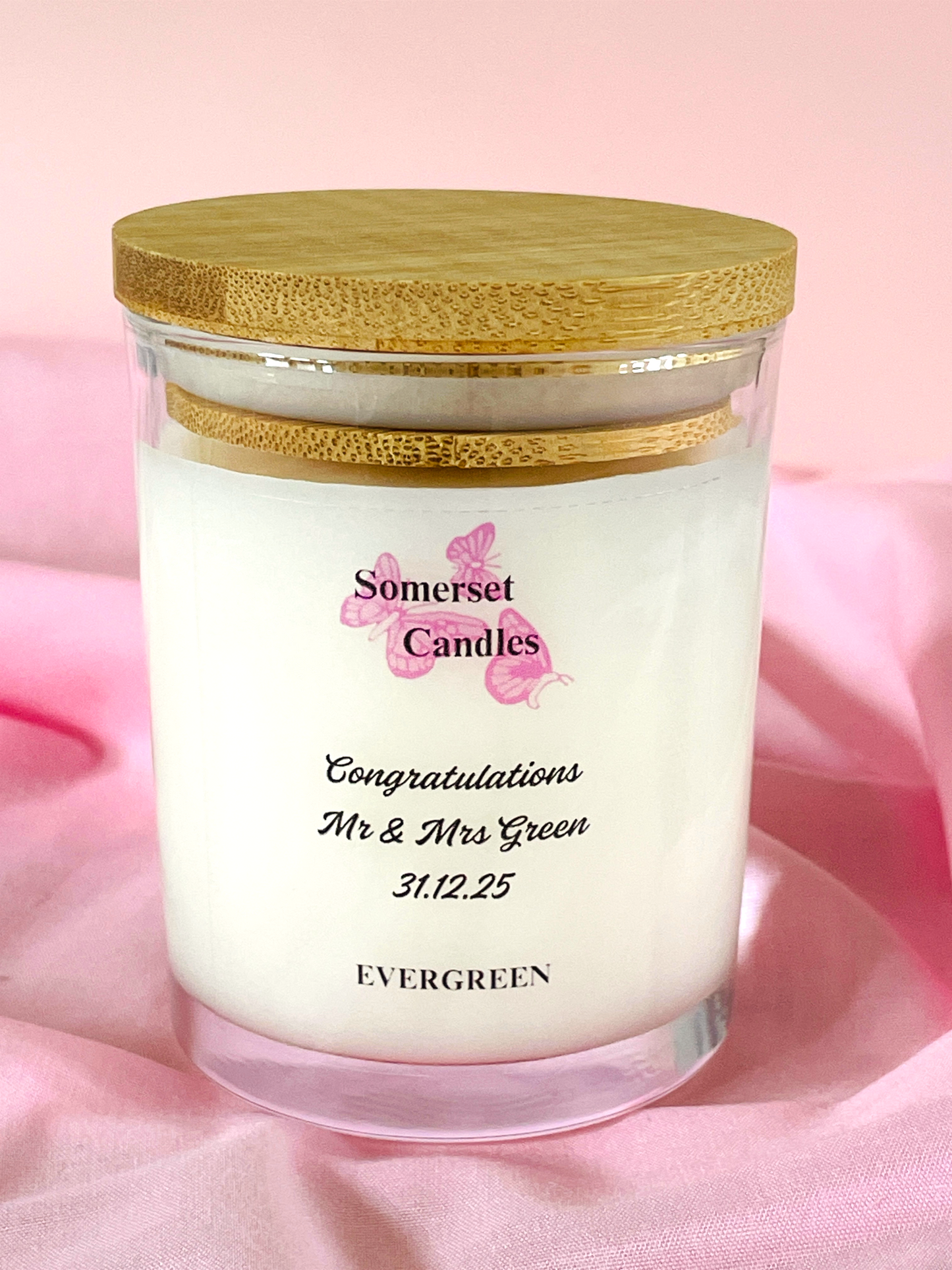 Candle in a glass jar with a wooden lid on a pink fabric background, featuring 'Somerset Candles' branding.