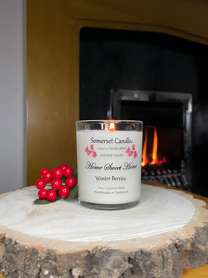 Home Sweet Home Winter Berries soy candle is displayed on a wooden board with berries in front of a lit fire The candle is white in colour with bright red butterflies fluttering above the scent name and fitted with a lovely bamboo lid.