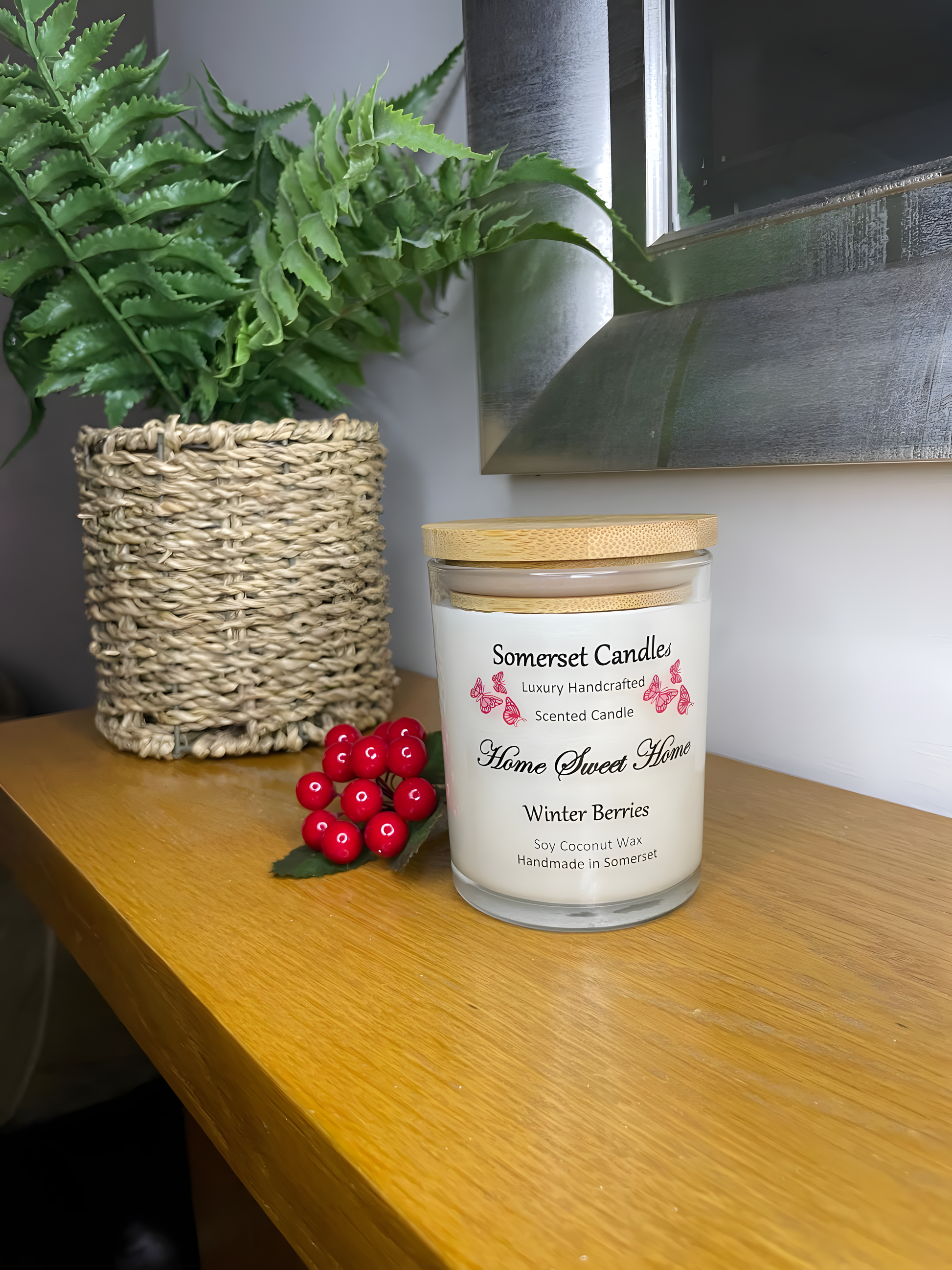 Home Sweet Home Winter Berries soy candle is displayed on a the mantelpiece with berries . The candle is white in colour with bright red butterflies fluttering above the scent name and fitted with a lovely bamboo lid.