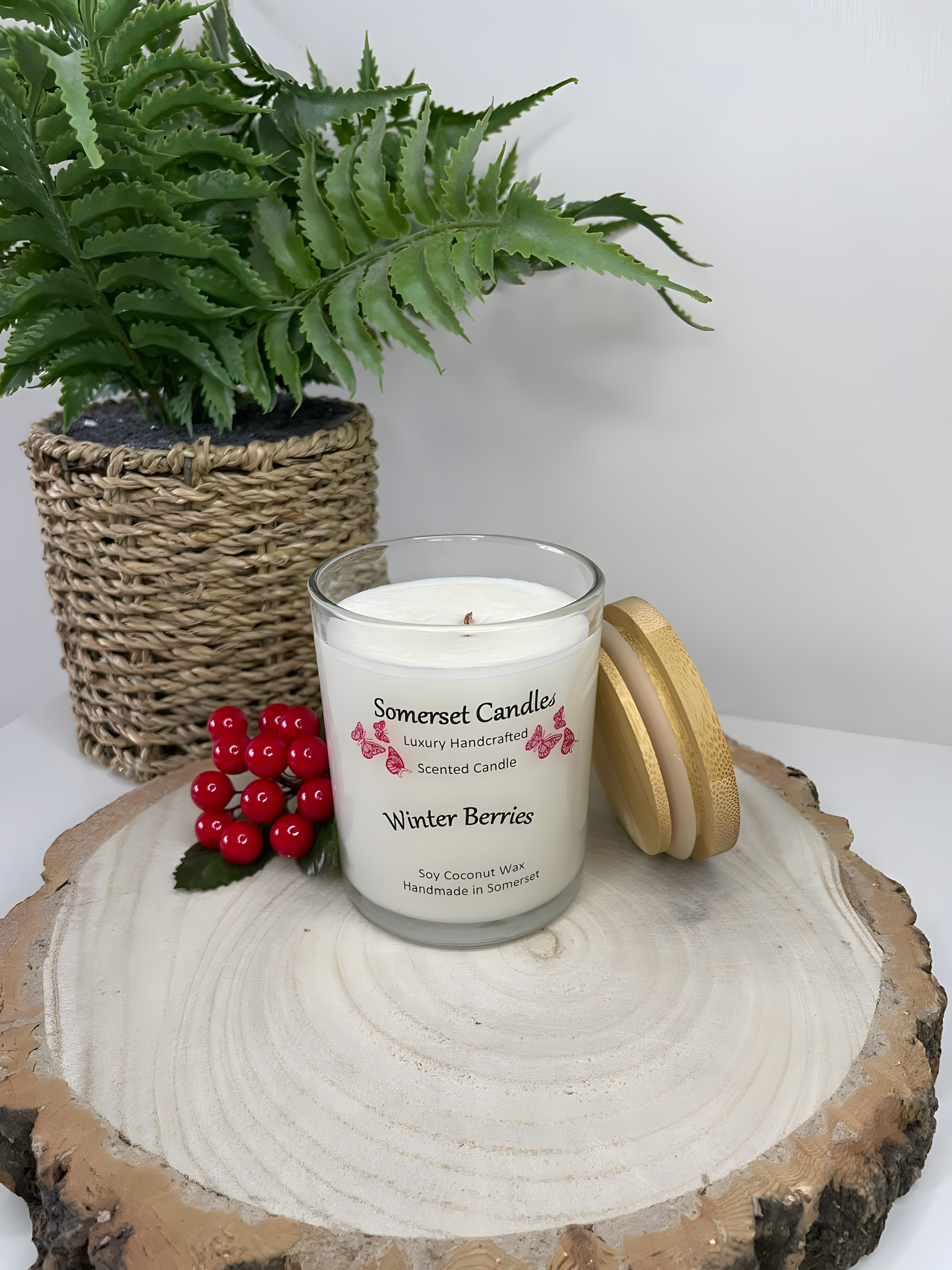 Winter Berries Scented Soy Candle displayed on a wooden board with berries and a plant behind. The candle is white in colour with bright red butterflies fluttering above the scent name. A bamboo lid is lent against the right side of the candle.