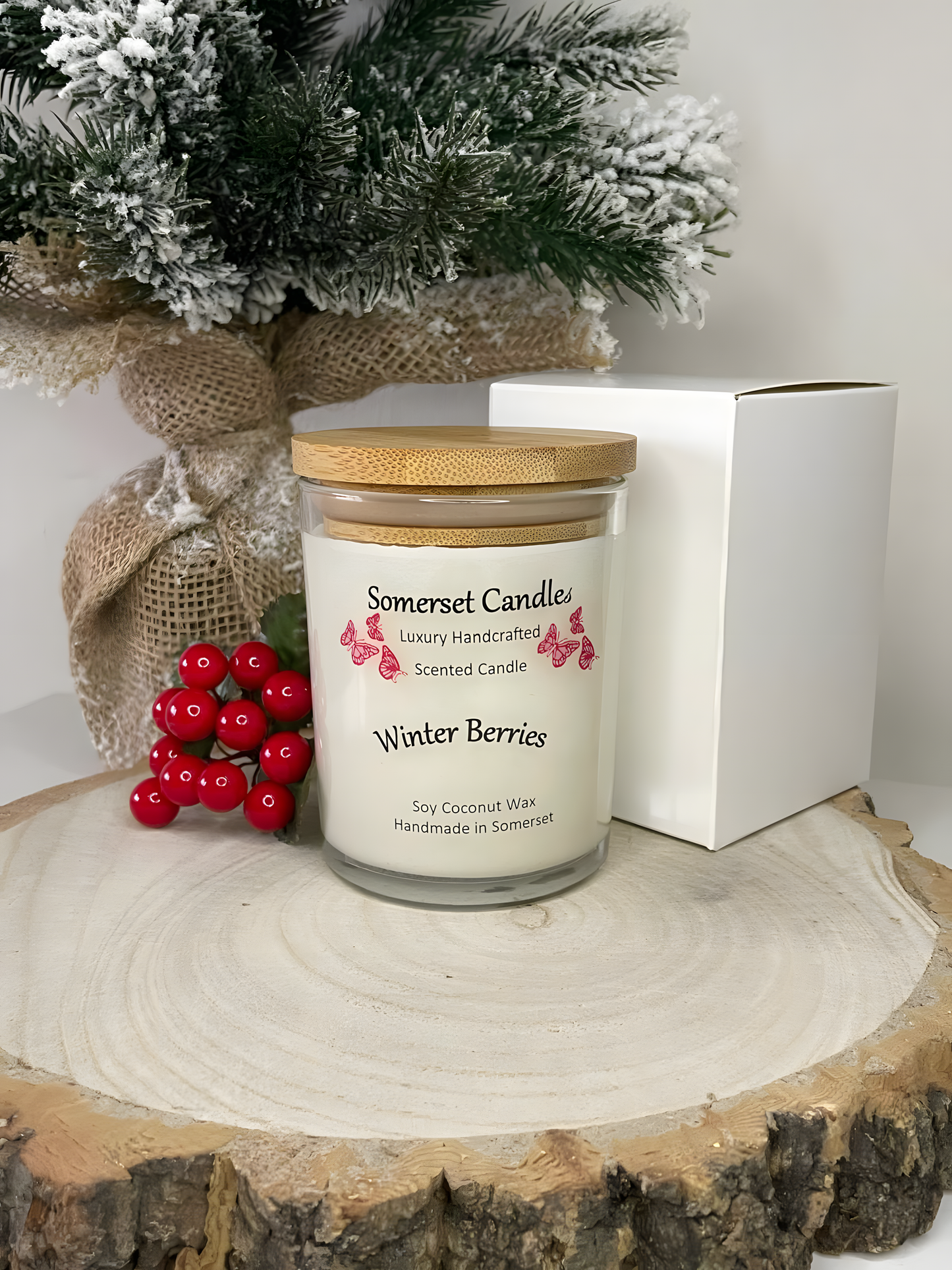 Winter Berries Scented soy candle displayed on a wooden board with berries and gift box behind. The candle is white in colour with bright red butterflies fluttering above the scent name and fitted with a lovely bamboo lid.